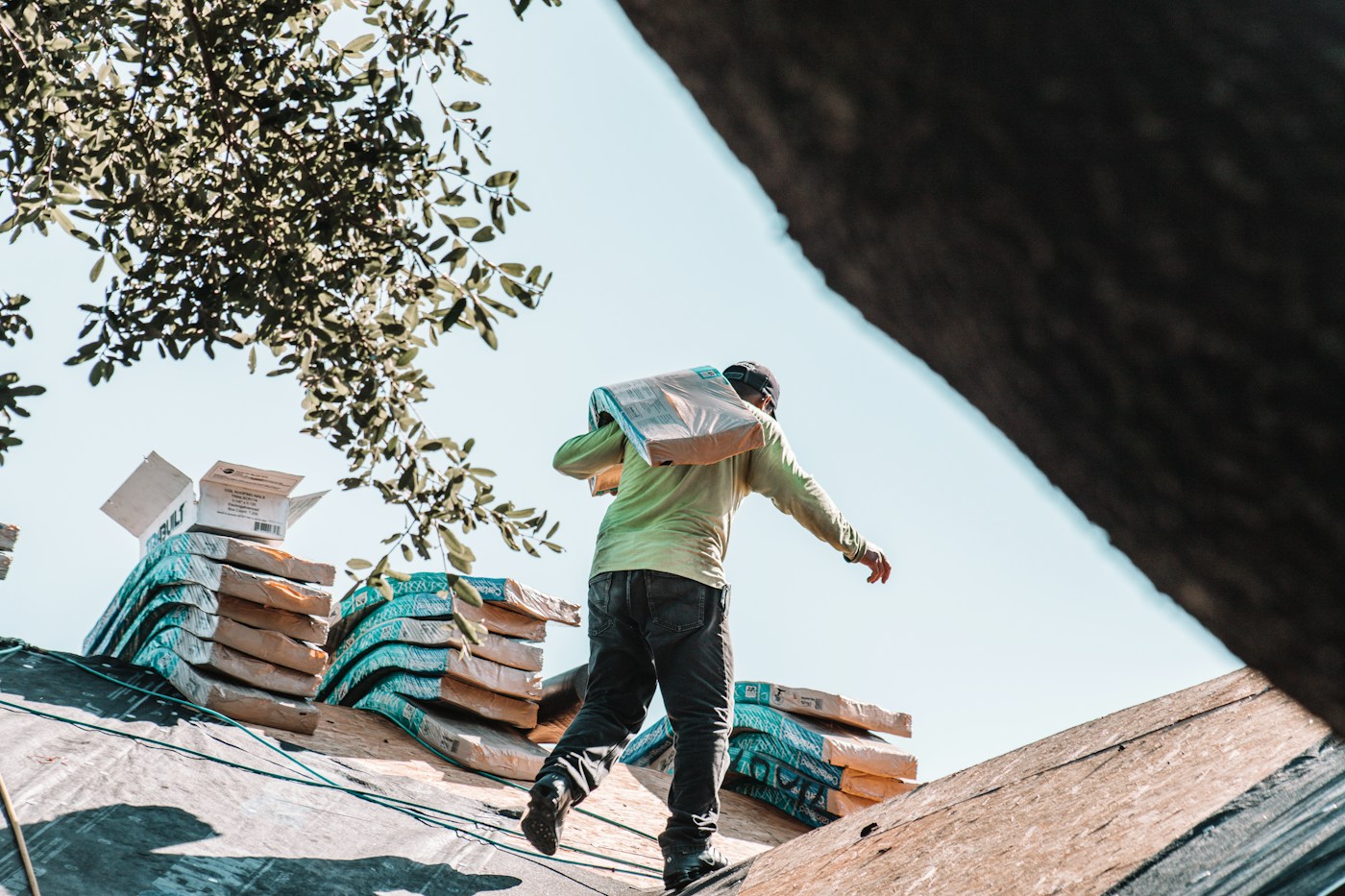 A roofer carries a bundle of shingles across a roof deck
